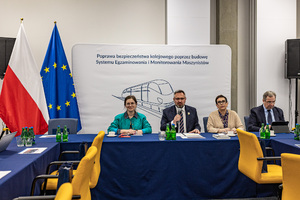 Sala konferencyjna, dwie kobiety oraz dwóch mężczyzn siedzi przy stole. W tle widać flagi Polski oraz Unii Europejskiej.