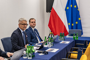 Dwóch mężczyzn Prezes Urzędu Transportu Kolejowego dr inż. Ignacy Góra oraz Piotr Szczepaniak Zastępca Dyrektora w  Departamencie Planowania i Nadzoru. Mężczyźni ubrani są w ciemne garnitury siedzą przy stole. Przed nimi znajdują się laptopy. W tle widać flagi Polski i Unii Europejskiej