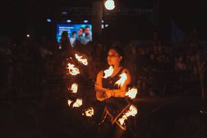 Na zdjęciu kobieta trzymająca stelaż z podpalonymi elementami którymi macha podczas pokazu fire show. Zdjęcie zrobione jest wieczorem