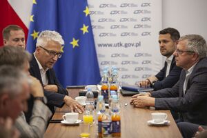 Mężczyźni siedzący po obu stronach brązowego stołu. Na środku napoje w butelkach oraz białe filiżanki. W tle flaga Polski, UE oraz logotypy UTK. Prezes UTK jest w trakcie wypowiedzi. Na wprost jest Prezydent Związku Zawodowego Maszynistów Kolejowych w Polsce.