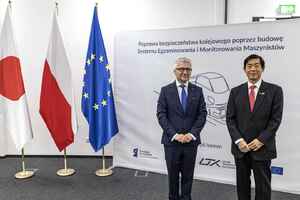 Na środku Ambasador Japonii w Polsce Miyajima Akio oraz Prezes UTK Ignacy Góra. W tle flaga Japonii, flaga Polski oraz flaga Unii Europejskiej. Mężczyźni mają garnitury z krawatami. Na podłodze szara wykładzina.