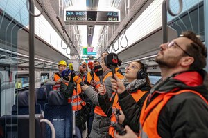 zdjęcie wnętrza pociągu, na środku stoi kilka osób w pomarańczowych kamizelkach odblaskowych z mikrofonami zwróconymi w kierunku sufitu pociągu nagrywają komunikaty głosowe nadawane z głośników. Wnętrze pociągu jest jasne, fotele granatowe.
