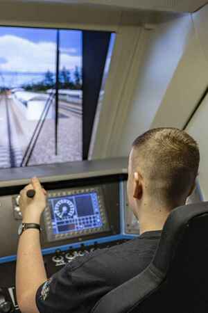 Wnętrze symulatora. Na fotelu siedzi mężczyzna przed nim widok z ekranu symulatora a na nim wyświetlana jest trasa jazdy pociągu.